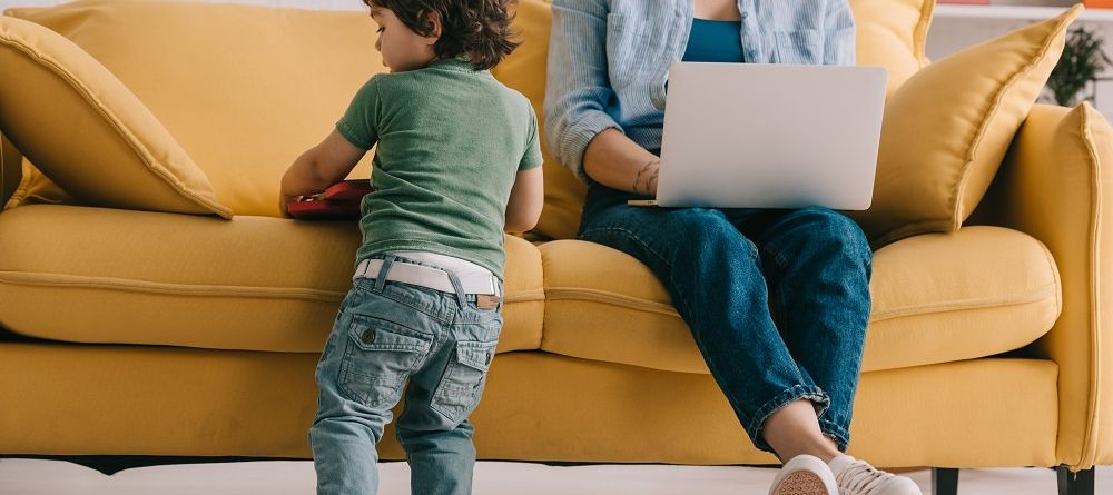 kid standing near sofa while mother working with laptop