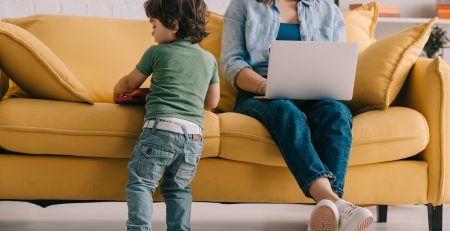 kid standing near sofa while mother working with laptop